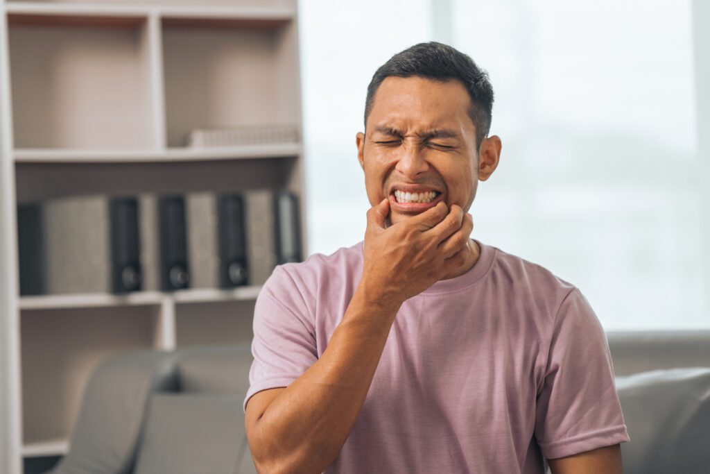 When Should I Go to an Emergency Dentist vs. the ER? - Viera Dental Arts | Dentist in Melbourne FL - Middle-aged Asian Indian man with gum disease, toothache, wisdom tooth and molar pain. sitting on the sofa