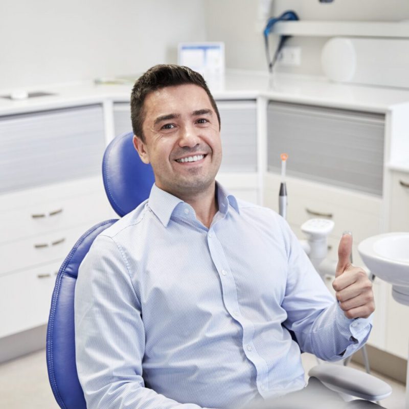 people, medicine, stomatology and health care concept - happy male patient sitting on dental chair and showing thumbs up at clinic office