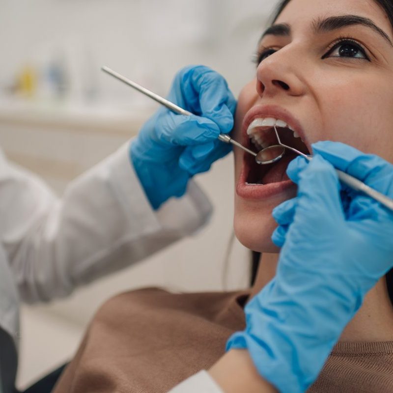 Dentist wearing blue gloves using dental mirror and probe examining patient's teeth during checkup appointment in modern dental clinic, providing professional oral healthcare