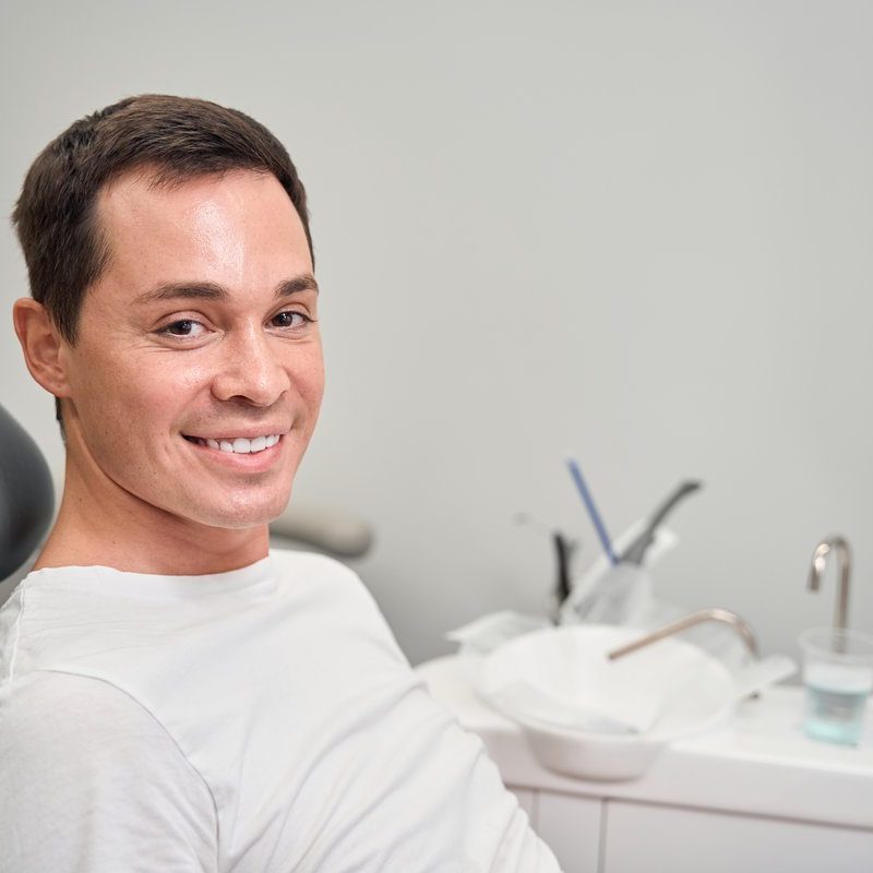 Positive handsome man sitting in dental chair and looking at camera with smile, visiting dentist for routine teeth health check-up, feeling free to smile, close up