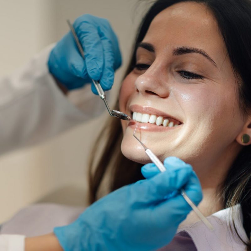 Dentist wearing blue gloves examining teeth of smiling young woman during a thorough dental checkup in a bright, clean clinic, ensuring optimal oral health and hygiene