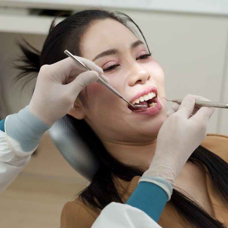 Asian female dental assistant performing dental cleaning using scaler and mirror on adult asian woman patient in dental chair smiling during oral hygiene treatment in modern dental clinic