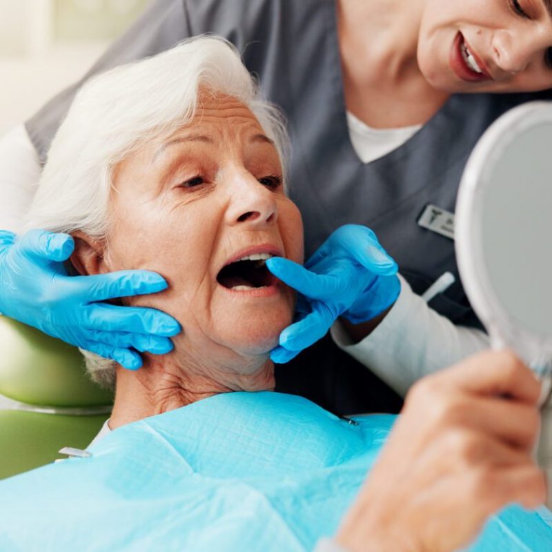 Dentist, senior woman and consulting with mirror for teeth cleaning, dental implant or oral hygiene. Inspection, healthcare or doctor with old patient in clinic for mouth exam or root canal treatment.