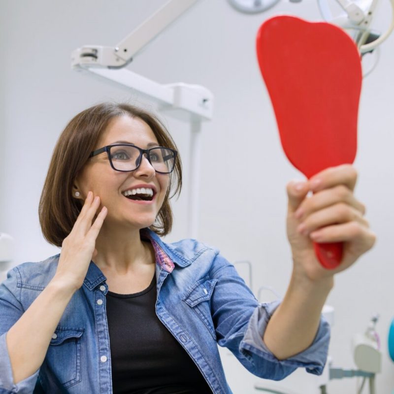 Woman patient looking in the mirror at the teeth, sitting in the dental chair. Healthcare, medical and dentistry concept.