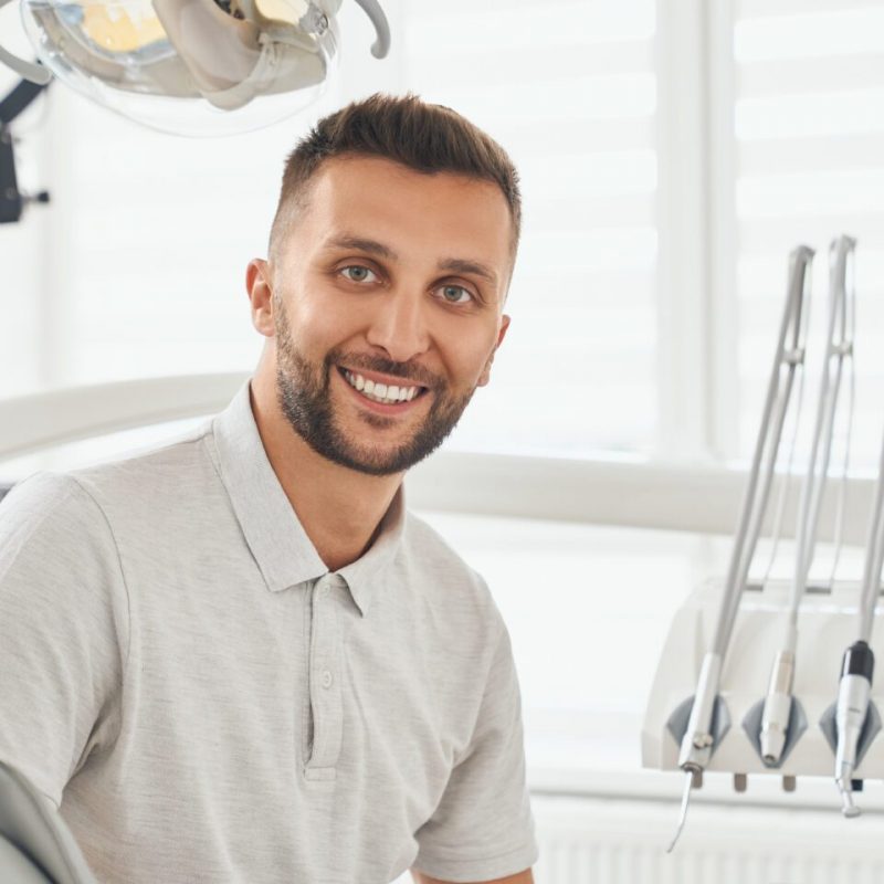 Portrait of happy bearded man in grey t-shirt sitting in dental chair, smiling and looking at camera. Young male patient looking satisfied after hygiene procedure at dental clinic