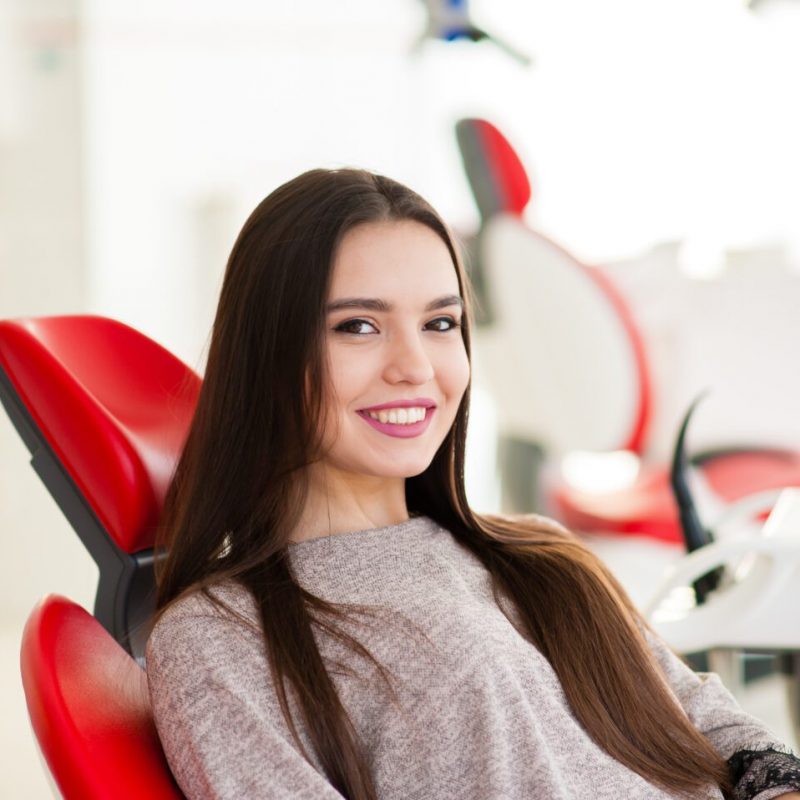 Young woman patient in checkered shirt with perfect straight white teeth waiting for dentist in dental chair and smiling relaxed, ready for a check-up. Beautiful woman smile