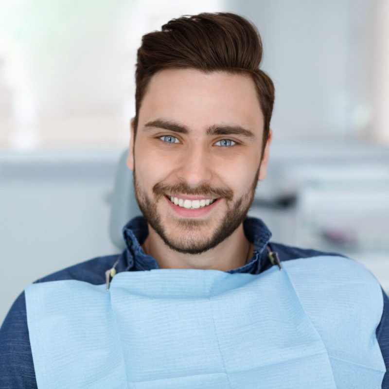 Cheerful bearde man patient sitting in dental chair and smiling at camera, copy space. Positive millennial guy showing his beautiful white smile after treatment at modern dental clinic