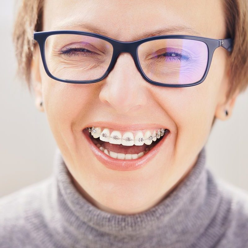 Stylish and beautiful young blonde woman in glasses and a gray sweater in braces and smiling on a white background, dentist orthodontist concept