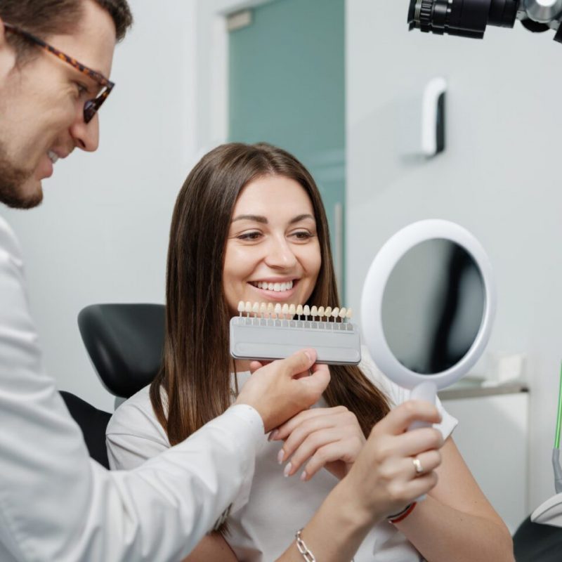 Young male dentist in white coat show veneers enamel color palette for beautiful patient woman sitting in a dental unit. Healthcare oral aesthetic treatment.