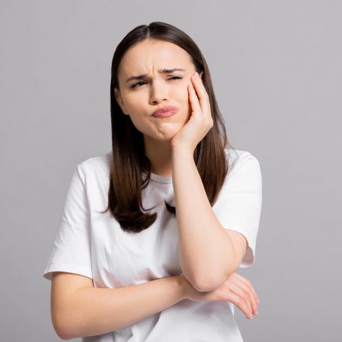 Crying tired sad woman suffers from painful toothache dental illness pointing on teeth standing on grey background in studio isolated wearing basic white t-shirt.