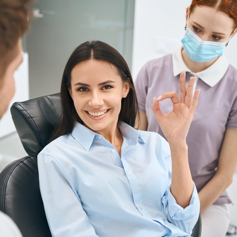 Smiling beautiful woman showing ok gesture sign sitting in orthodontic chair after painless and quality dental treatment, qualified dentist and nurse standing nearby