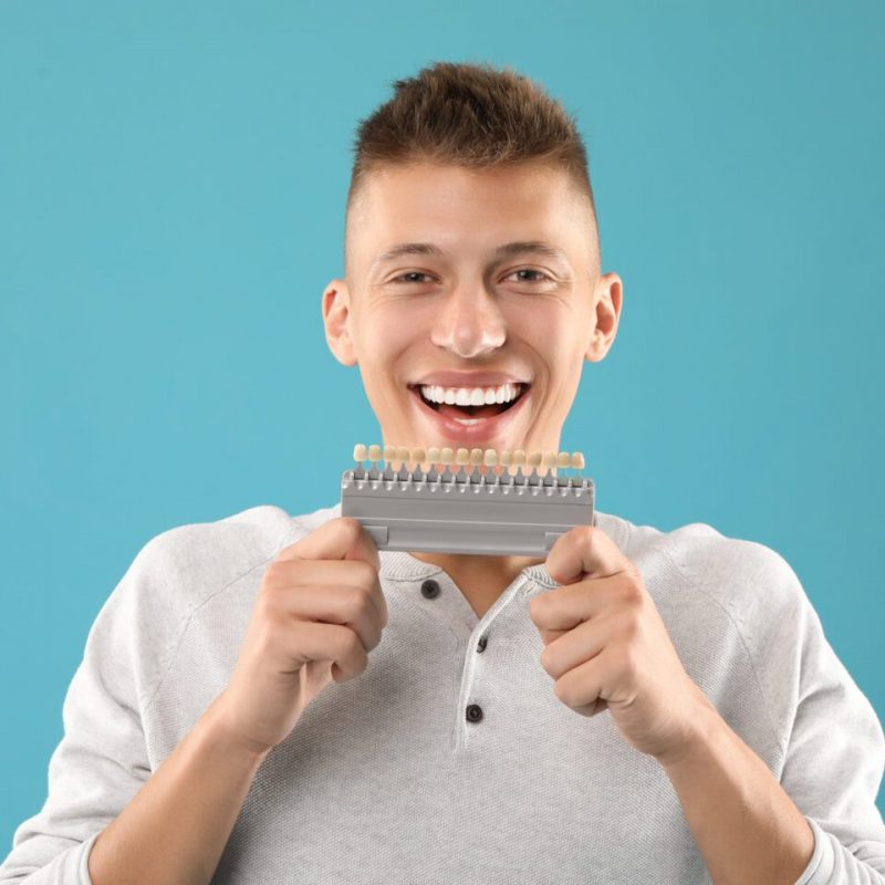 Happy young man with teeth color samples on light blue background. Dental veneers